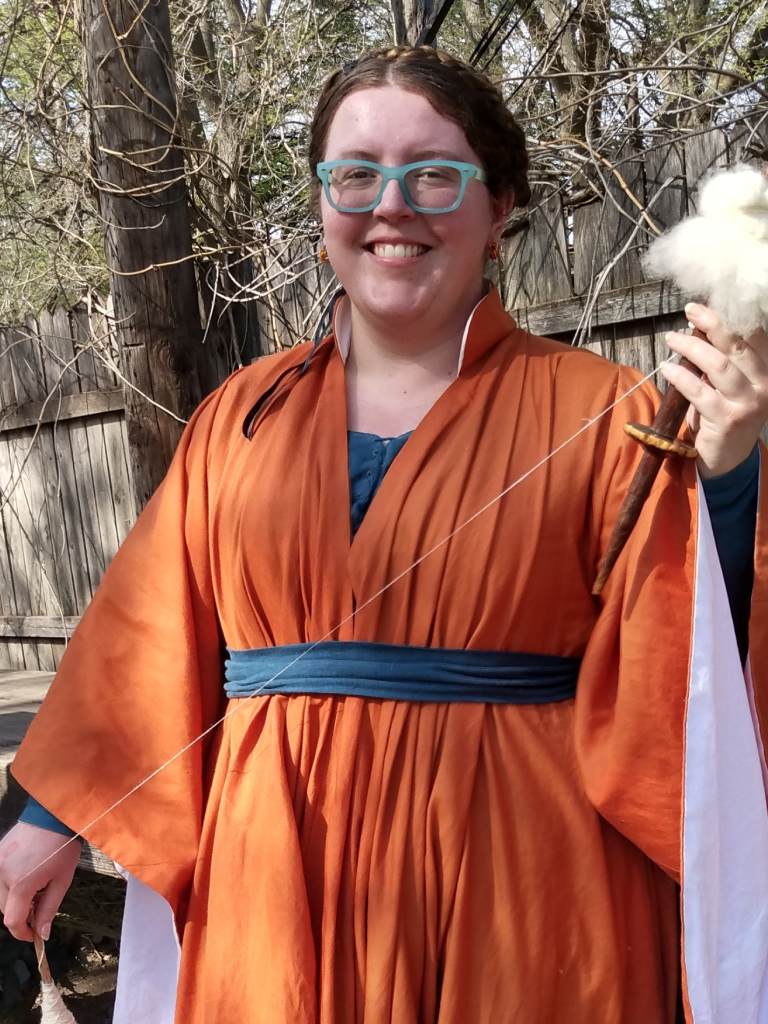 The Webminister wearing an orange flowing dress, holding a spindle and distaff